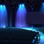 Moderno auditorio con iluminación azul y púrpura, escenario principal con pantallas LED mostrando gráficos financieros avanzados