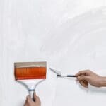 Primer plano de un pincel aplicando pintura sobre una pared blanca en un entorno moderno.