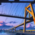 Puente moderno de acero al amanecer con ciudad de fondo, destacando ingeniería avanzada.