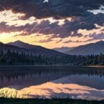 Vista panorámica de un lago con montañas y bosques al fondo iluminados por el sol.